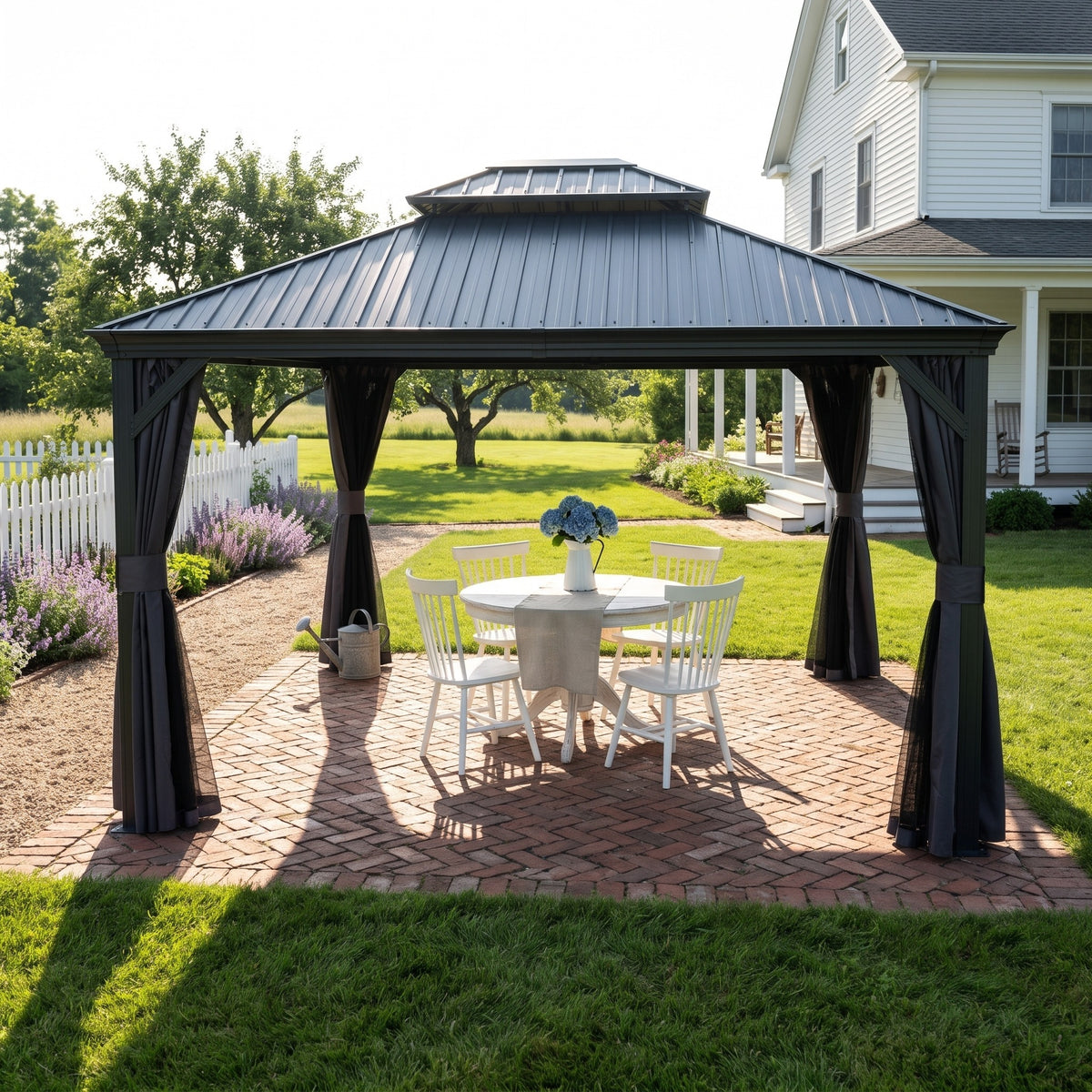 Dark gray hardtop gazebo with tied back curtains over a white table and chairs on a brick patio beside a farmhouse