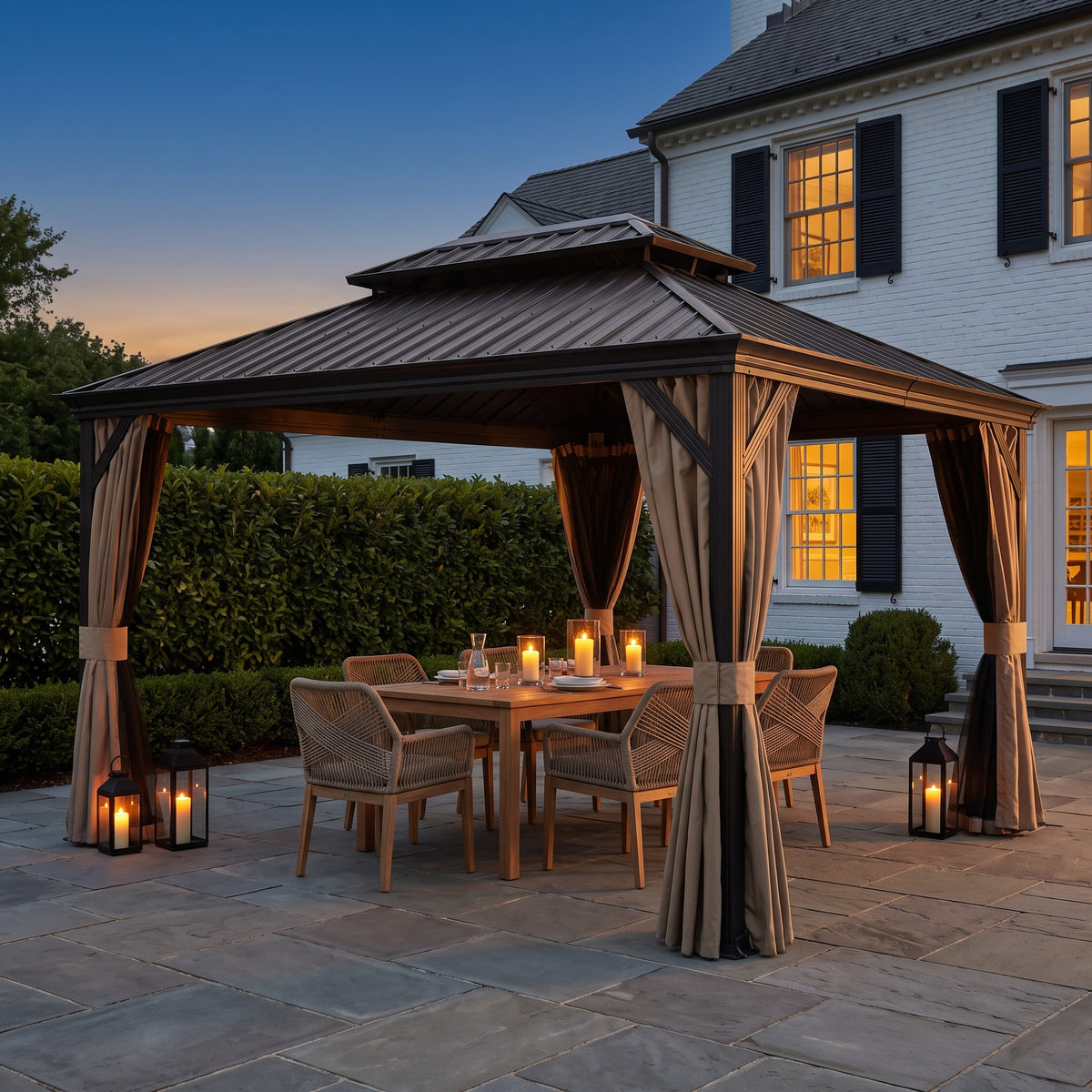 Brown 10x14 hardtop gazebo with tied back curtains over a teak dining table and candles on a stone patio at dusk