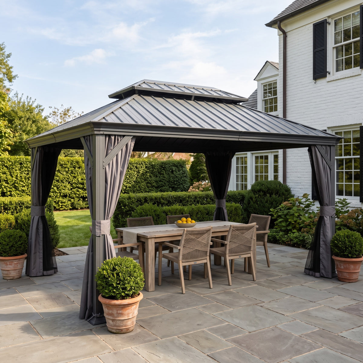 Gray double roof hardtop gazebo with tied back curtains over a wood dining table on a stone patio by a white house