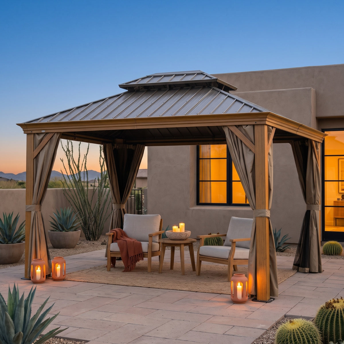 Wood grain aluminum 10x14 hardtop gazebo with tan curtains over two lounge chairs on a desert patio at dusk