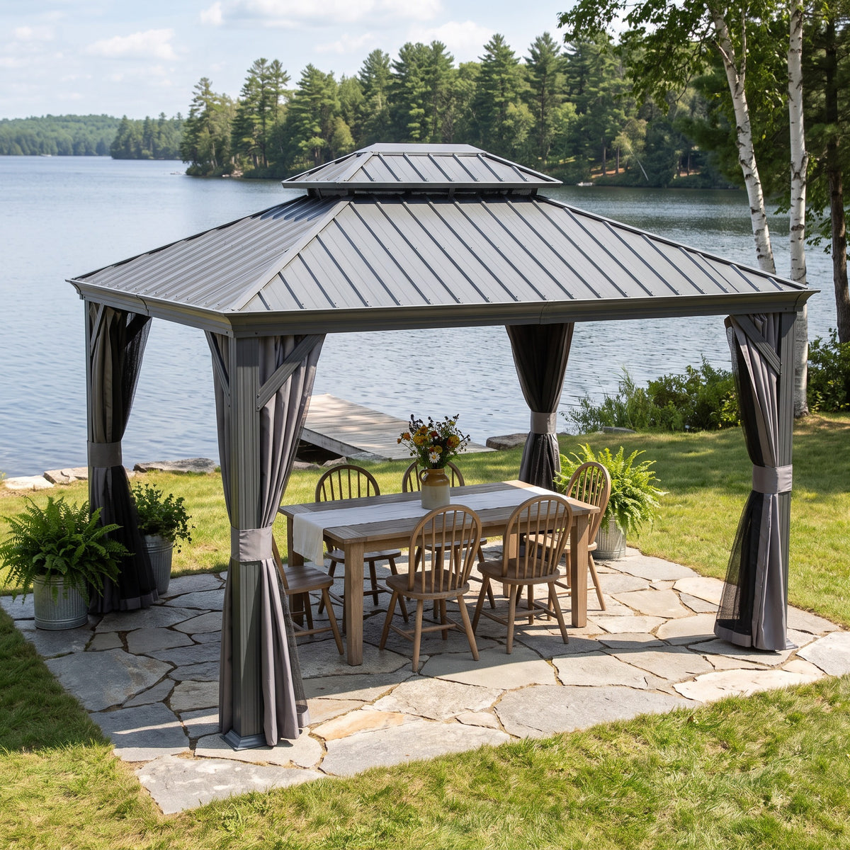 Gray steel double roof hardtop gazebo with curtains over a wood dining table on a stone patio by a lake