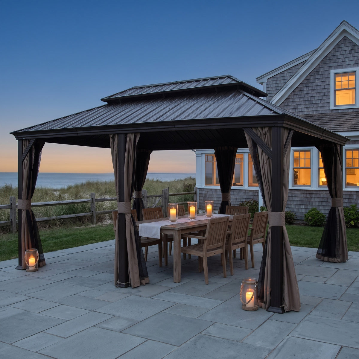 Brown 12x18 hardtop gazebo with tied back curtains over a teak dining table on a stone patio at dusk