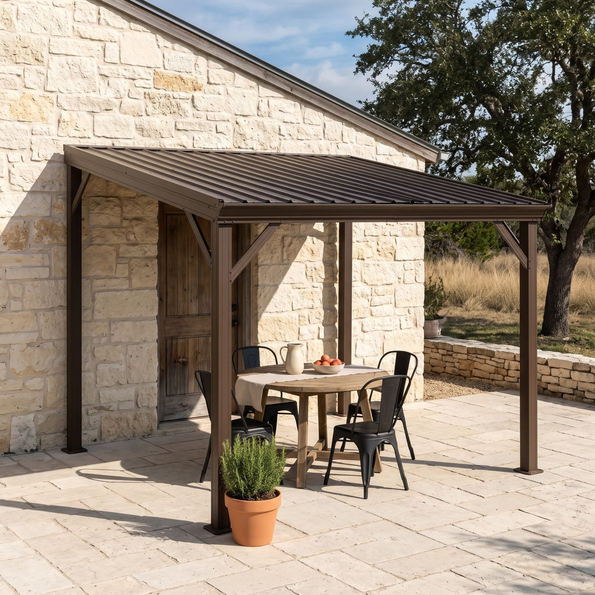 Brown metal lean-to hardtop gazebo over a round dining table beside a stone house wall in afternoon sun