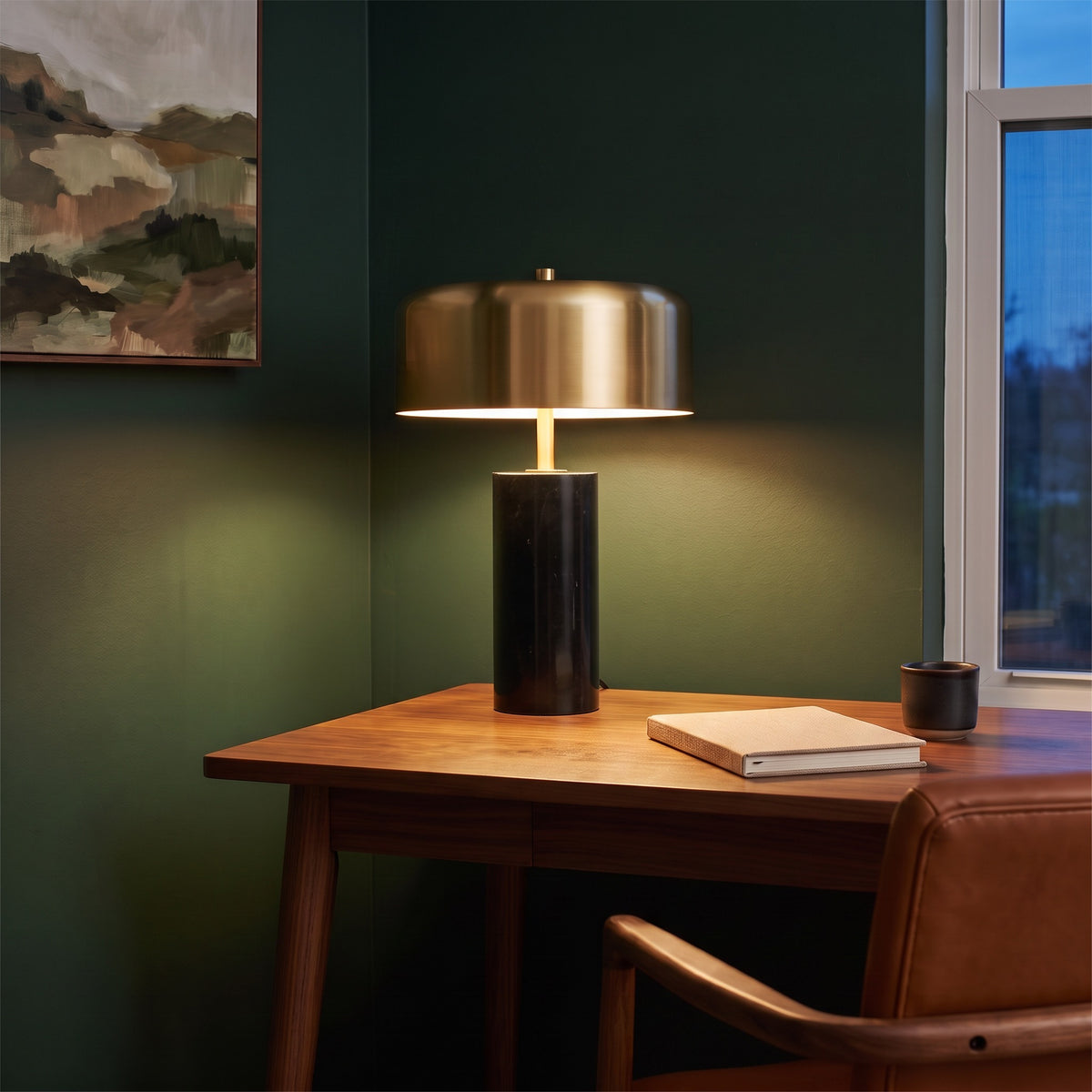 Black marble table lamp with gold drum shade, lit, on a walnut desk against a dark green wall at dusk