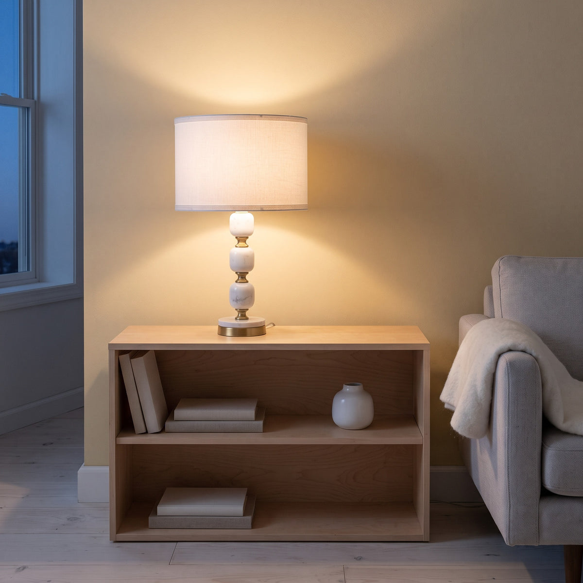 White marble table lamp with brass accents and white drum shade lit on a maple bookcase beside a sofa