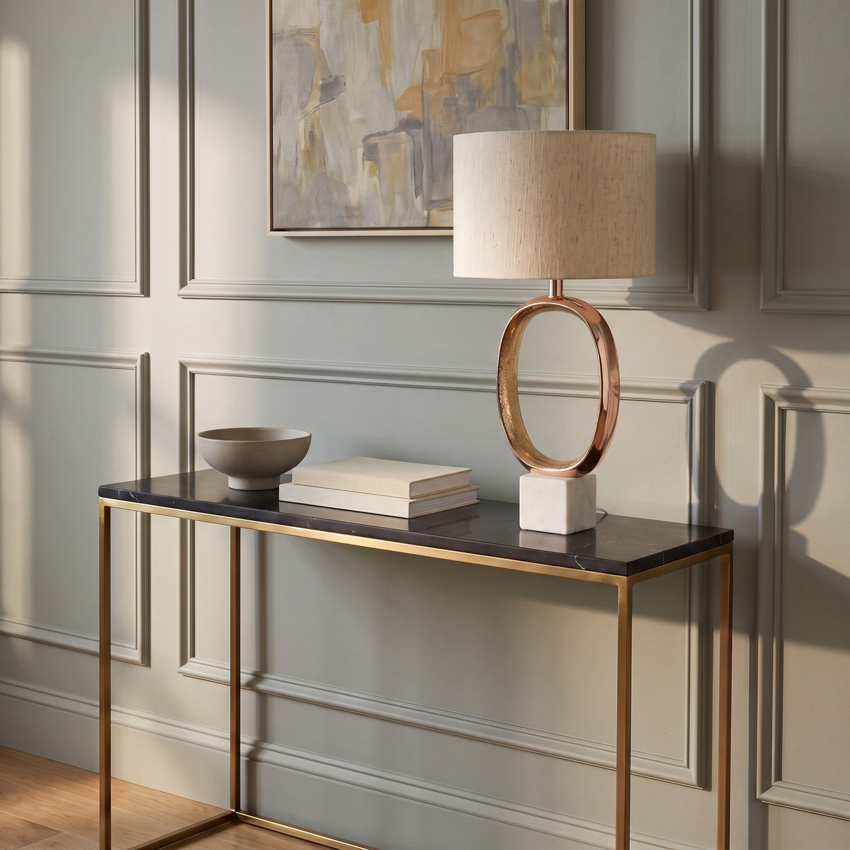 Rose gold oval cutout table lamp with linen drum shade and white marble base on a black marble console