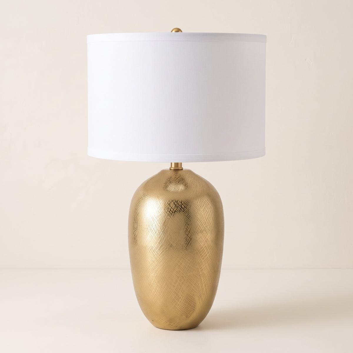 Gold aluminum table lamp with crosshatch texture and white drum shade on an ivory backdrop