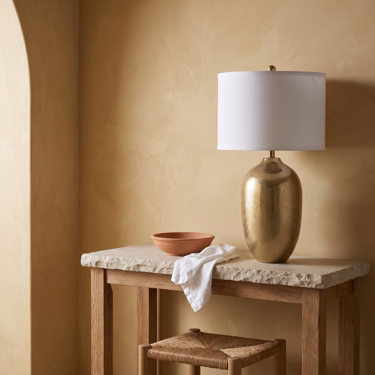 Gold etched urn table lamp with white drum shade on a stone top oak console against a plaster wall