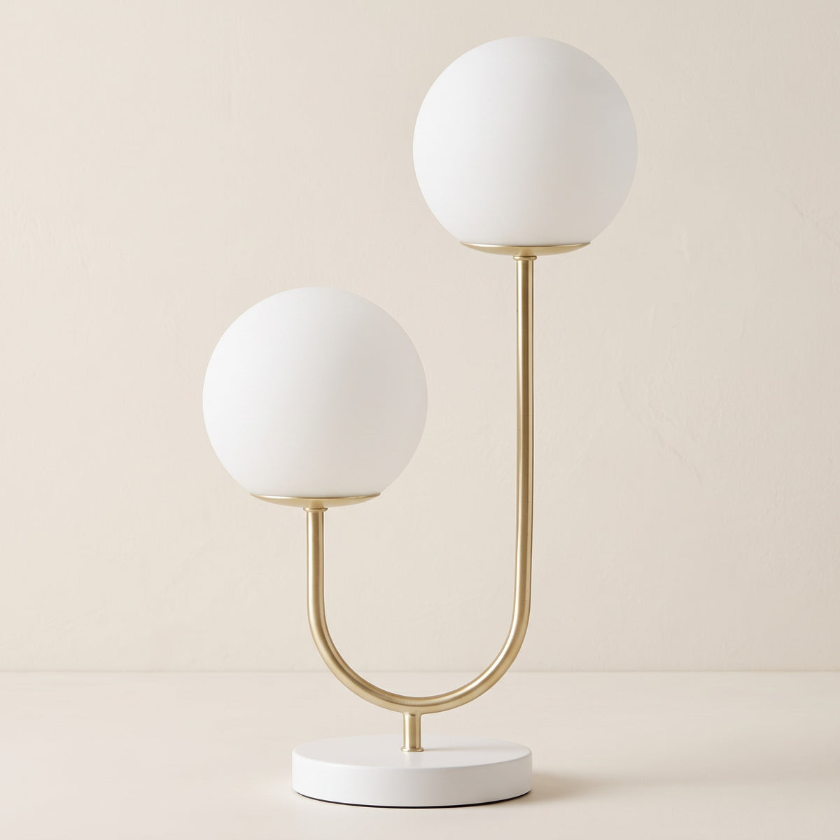 Gold and white table lamp with two white glass globe shades on a curved arm against a warm ivory backdrop