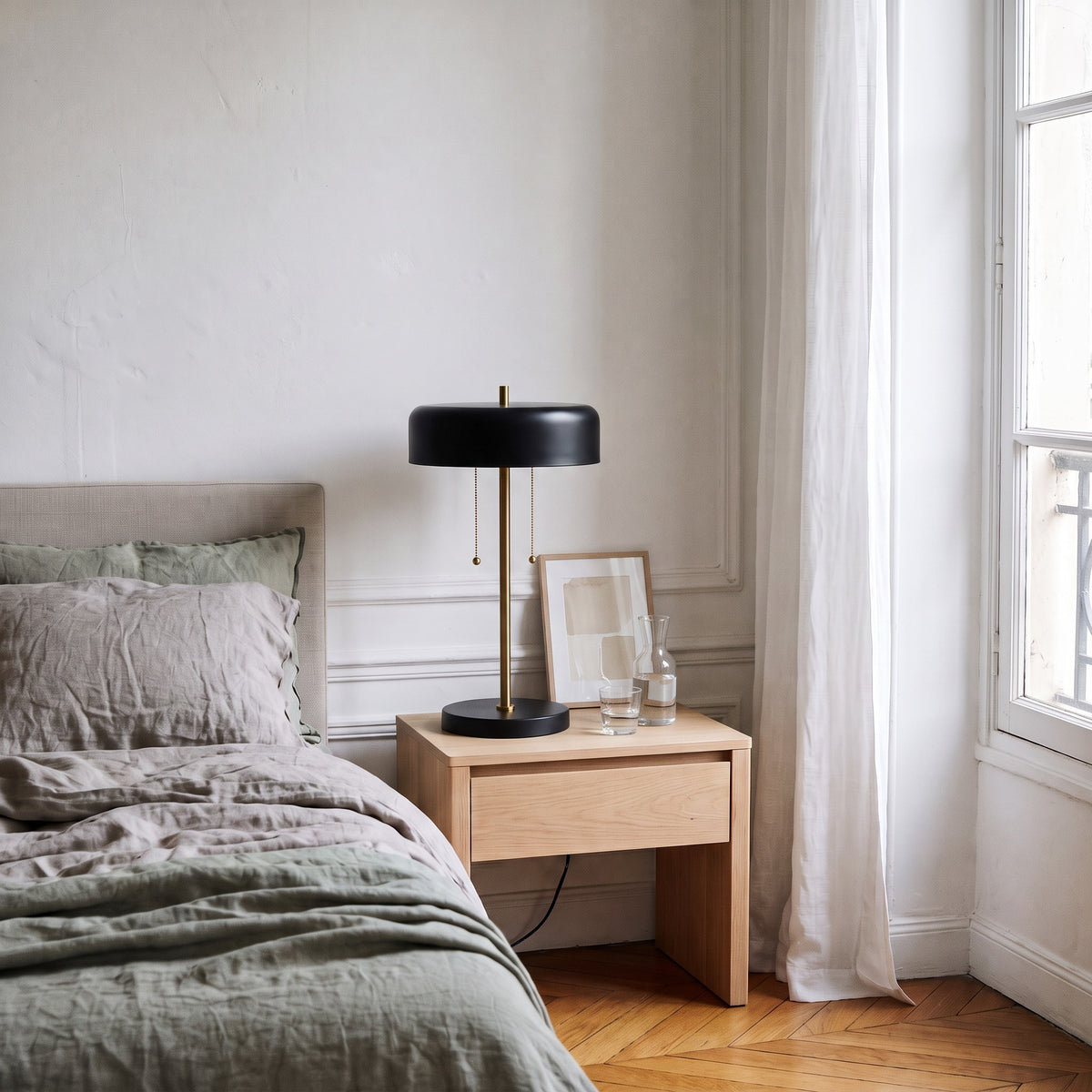 Black metal table lamp with brass stem and two pull chains on a light wood nightstand beside a linen bed