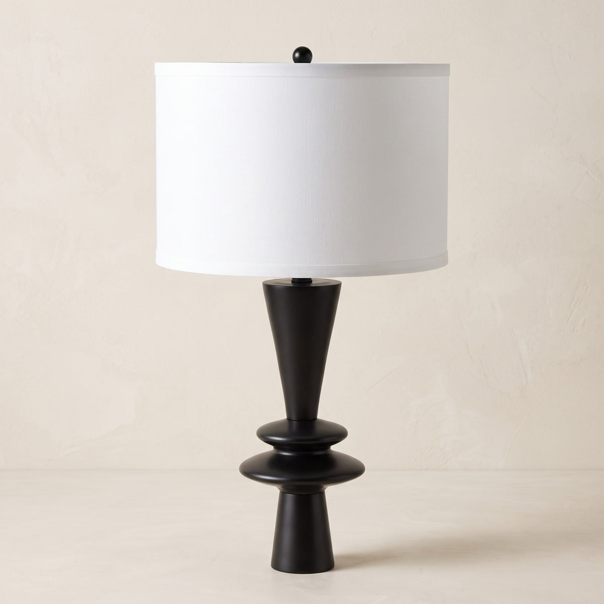 Black table lamp with stacked disc base and white fabric drum shade on an ivory plaster backdrop