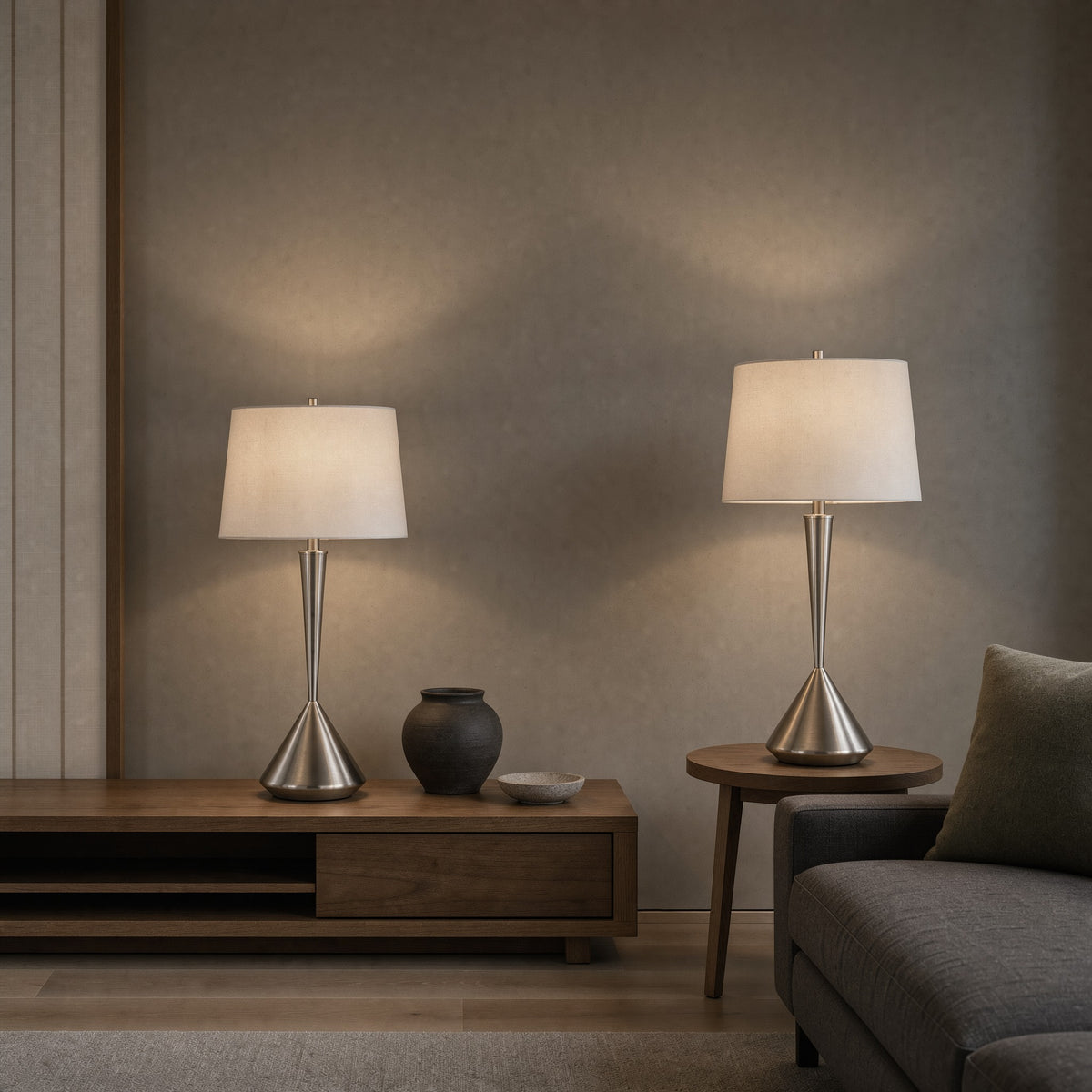 Two brushed nickel table lamps with white linen shades lit on a walnut console and side table beside a grey sofa