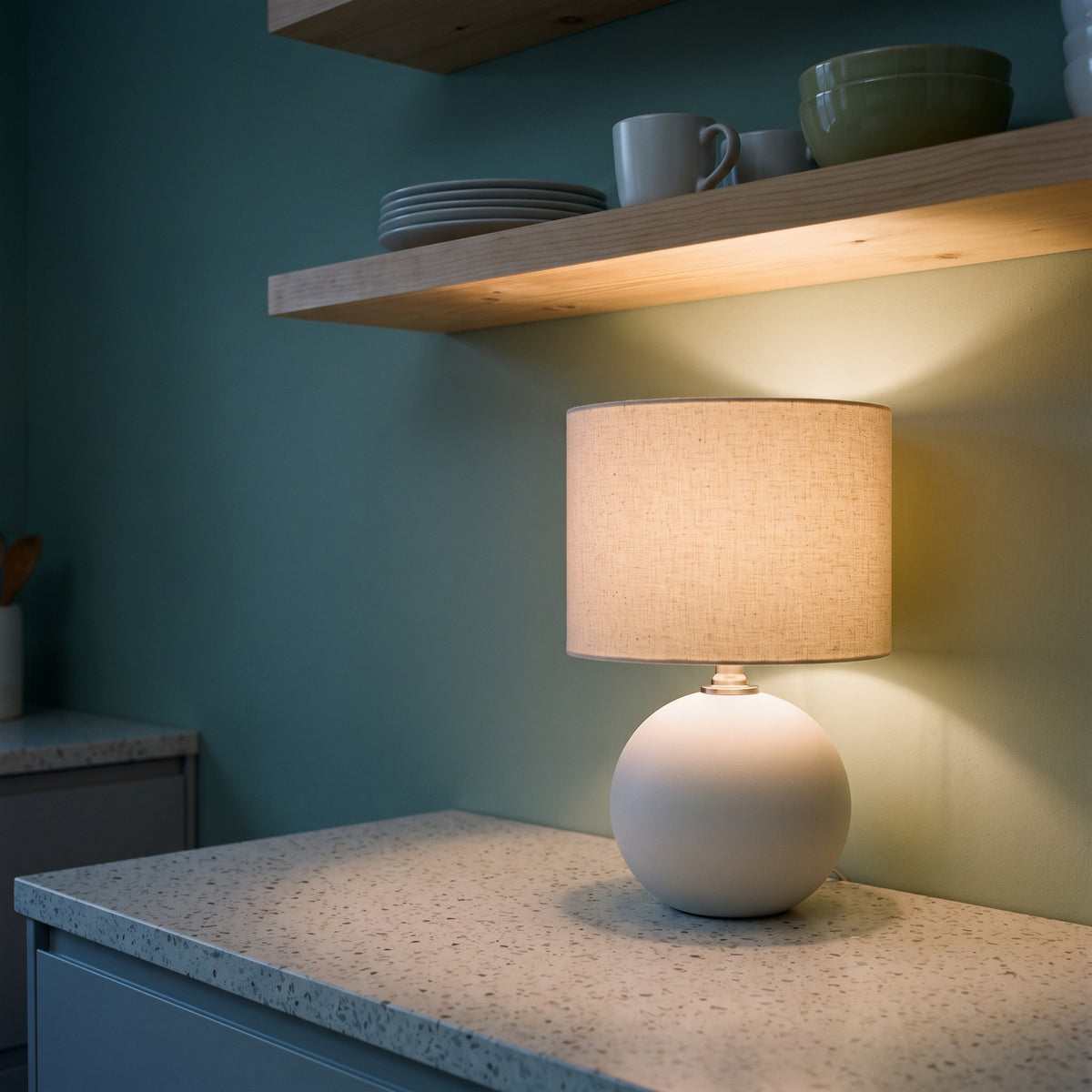 Cream ceramic sphere table lamp with oatmeal linen drum shade, lit, on a terrazzo kitchen counter below wood shelves