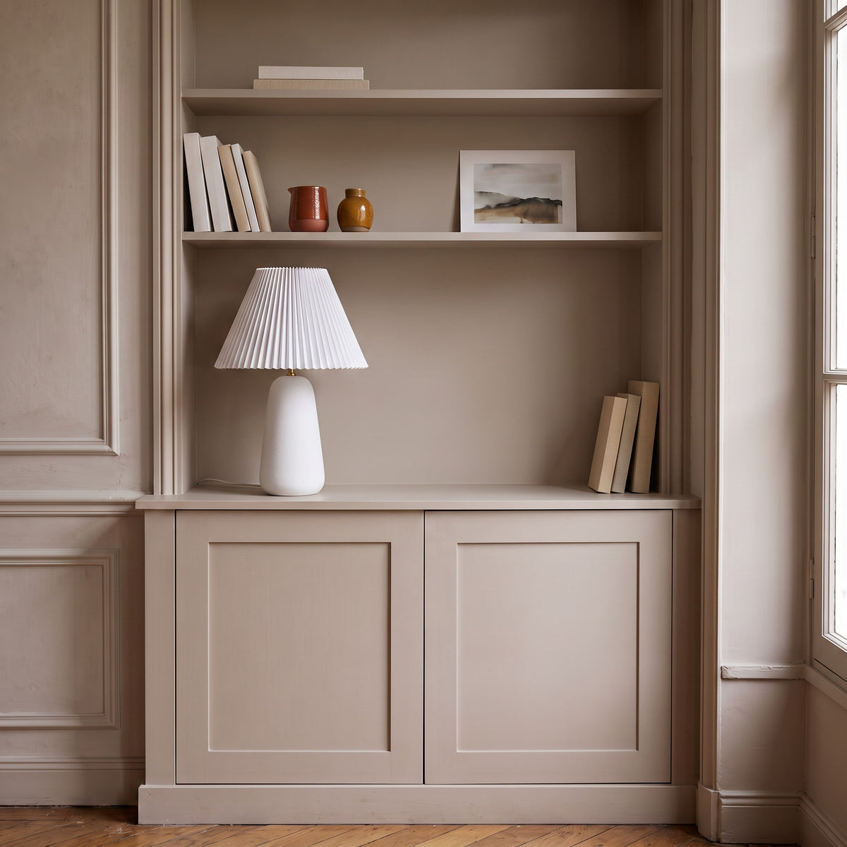 White textured ceramic table lamp with pleated white shade on a greige built-in cabinet beside shelves