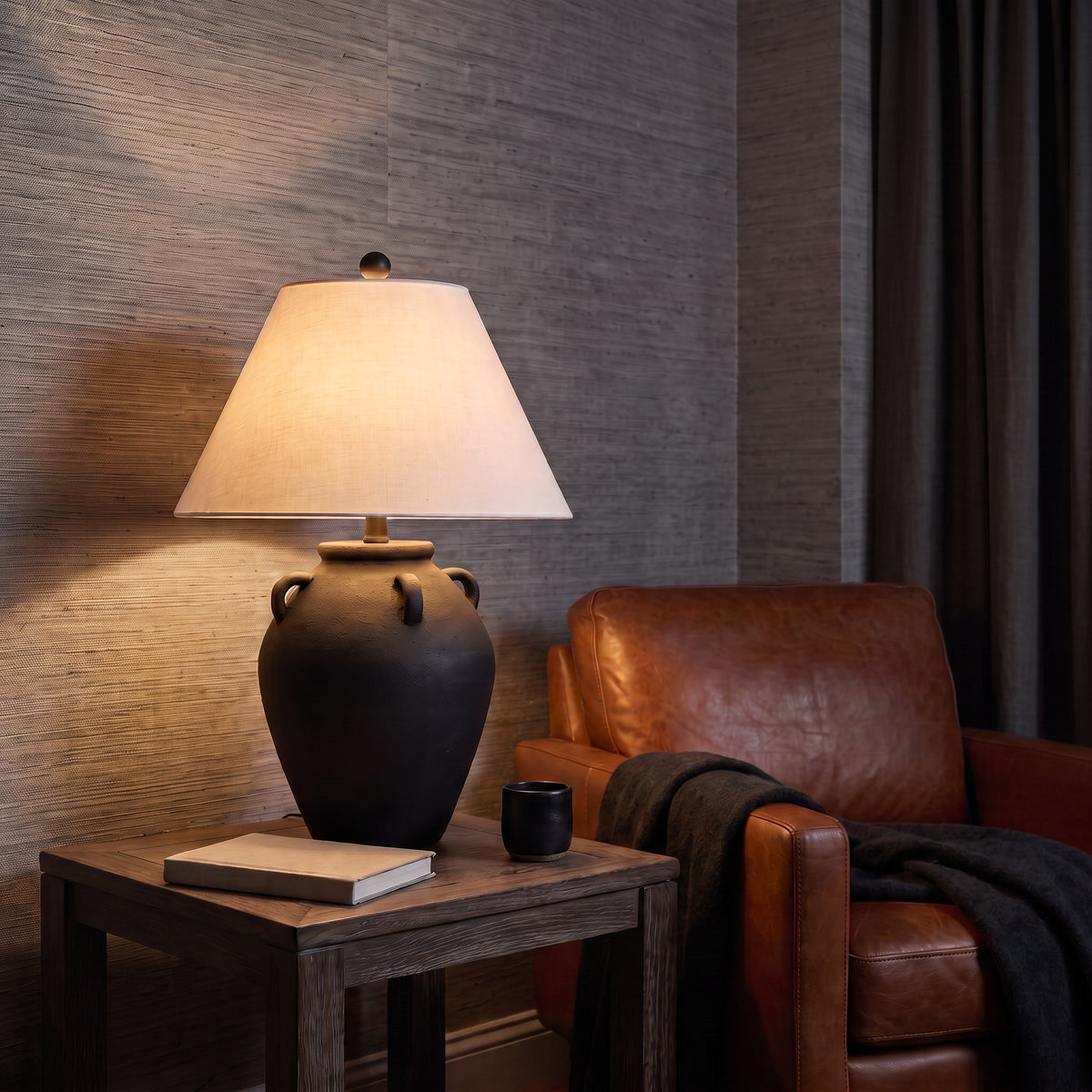 Black ceramic jug table lamp with white linen-look shade, lit on an oak side table beside a leather armchair