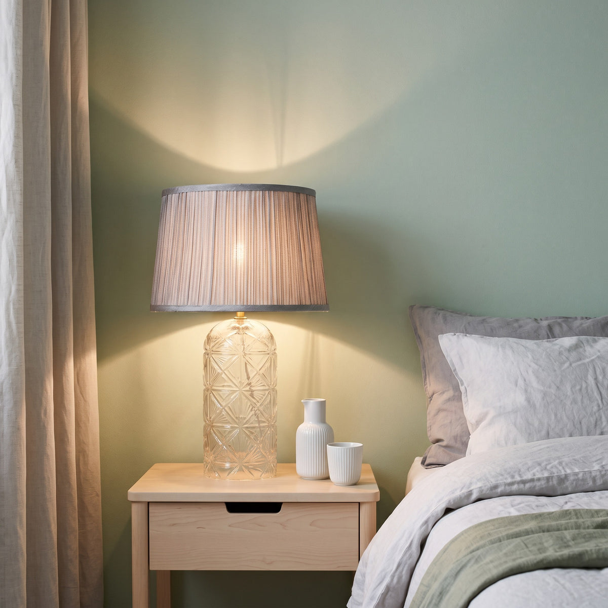Clear cut glass table lamp with pleated grey fabric shade, lit on a light wood nightstand beside a bed