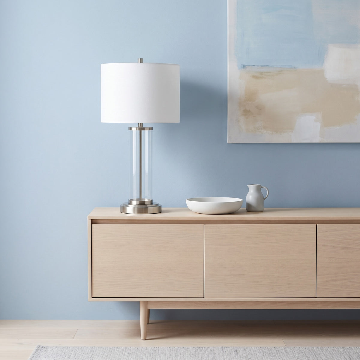 Clear glass table lamp with brushed nickel base and white drum shade on a light oak credenza against a blue wall