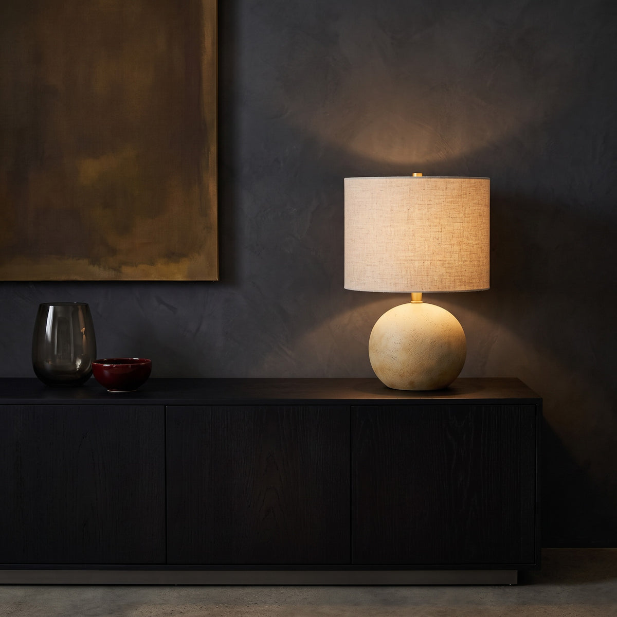 Beige concrete ball table lamp with linen drum shade, lit, on a black wood console against a dark plaster wall