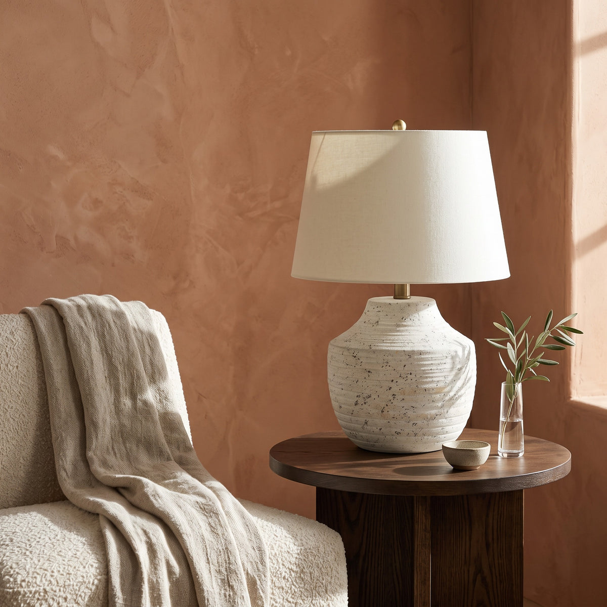 Cream concrete table lamp with ridged base and ivory linen shade on a dark wood side table by a boucle chair