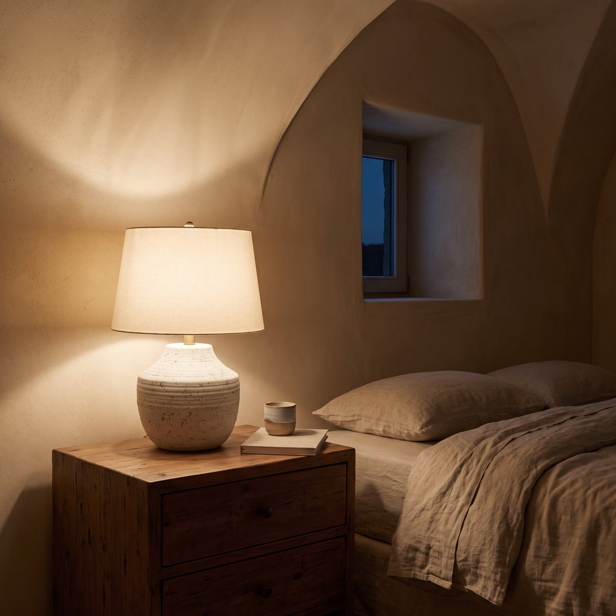 Cream concrete table lamp with ivory linen shade lit on a wood nightstand beside a linen bed