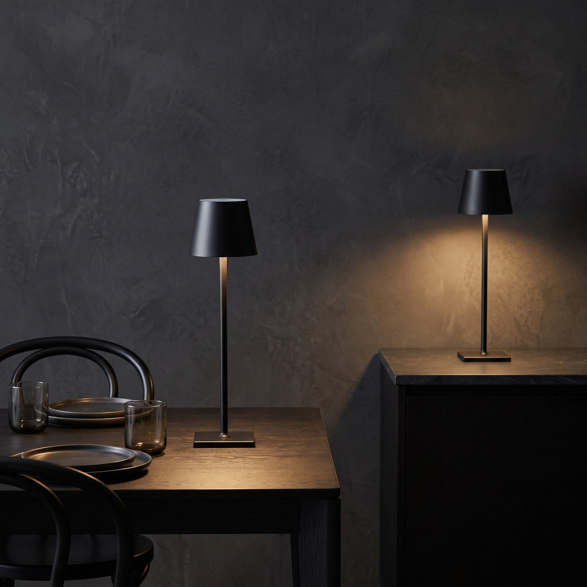 Two black cordless table lamps glowing on a dark wood dining table and a stone topped cabinet
