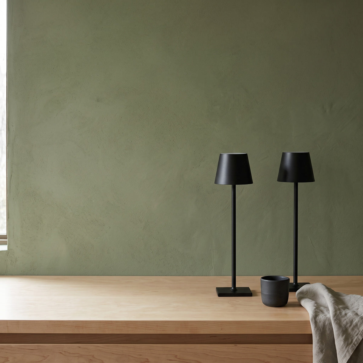 Two black cordless table lamps on a light wood sideboard with a dark ceramic cup and linen towel against a sage green wall
