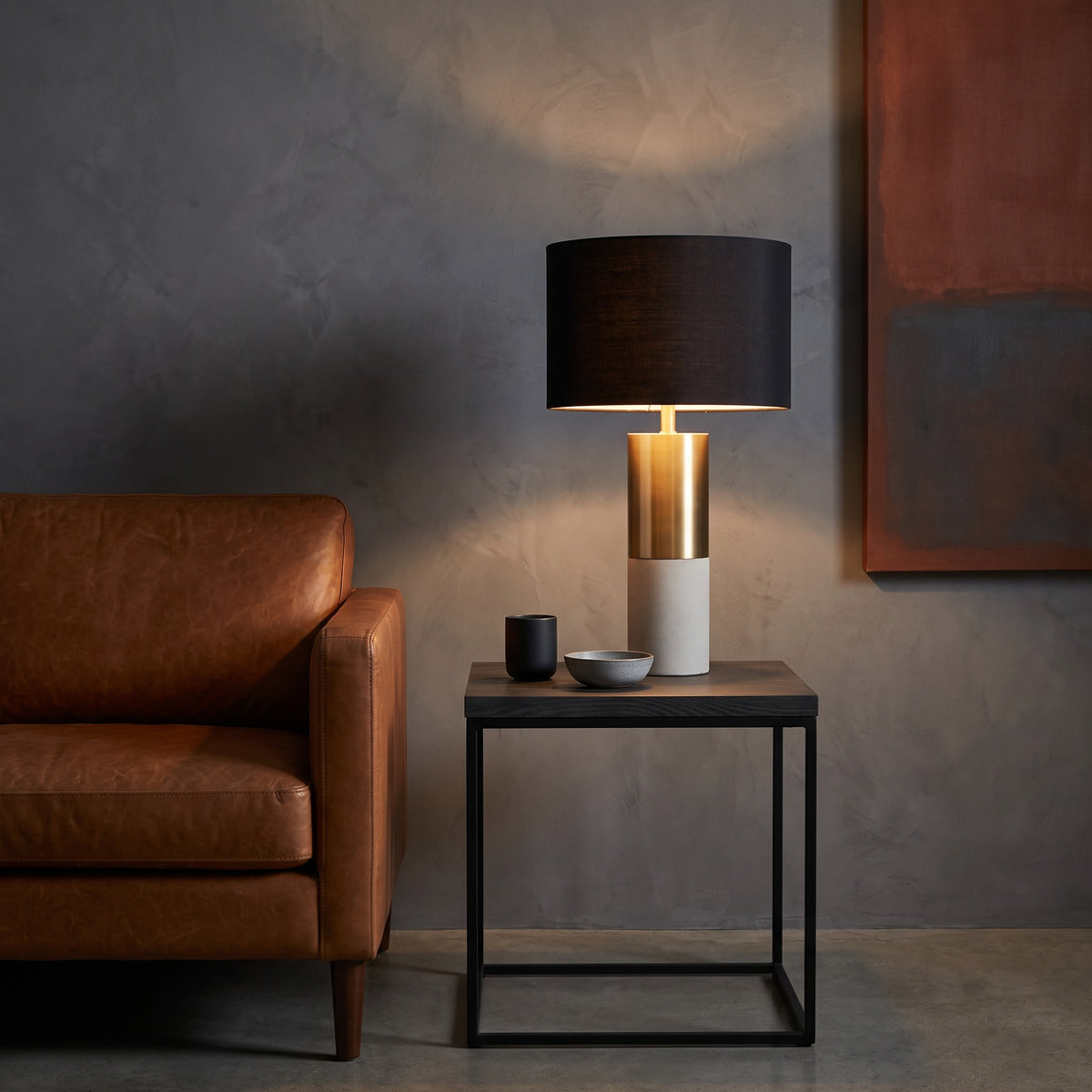 Black drum shade table lamp with brass and concrete base, lit on a dark side table beside a tan leather sofa
