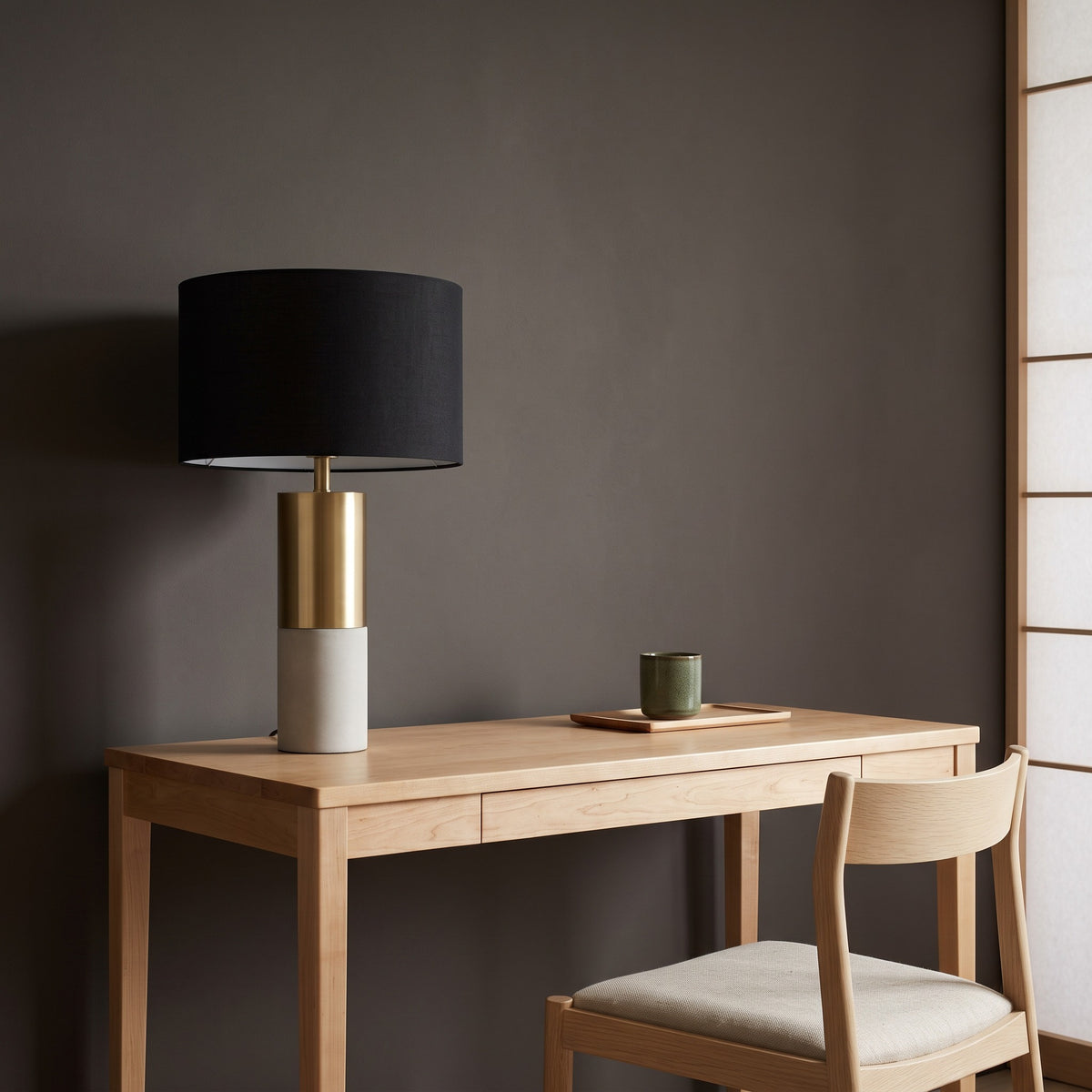 Black drum shade table lamp with brass and concrete column base on a light wood desk beside a wood chair