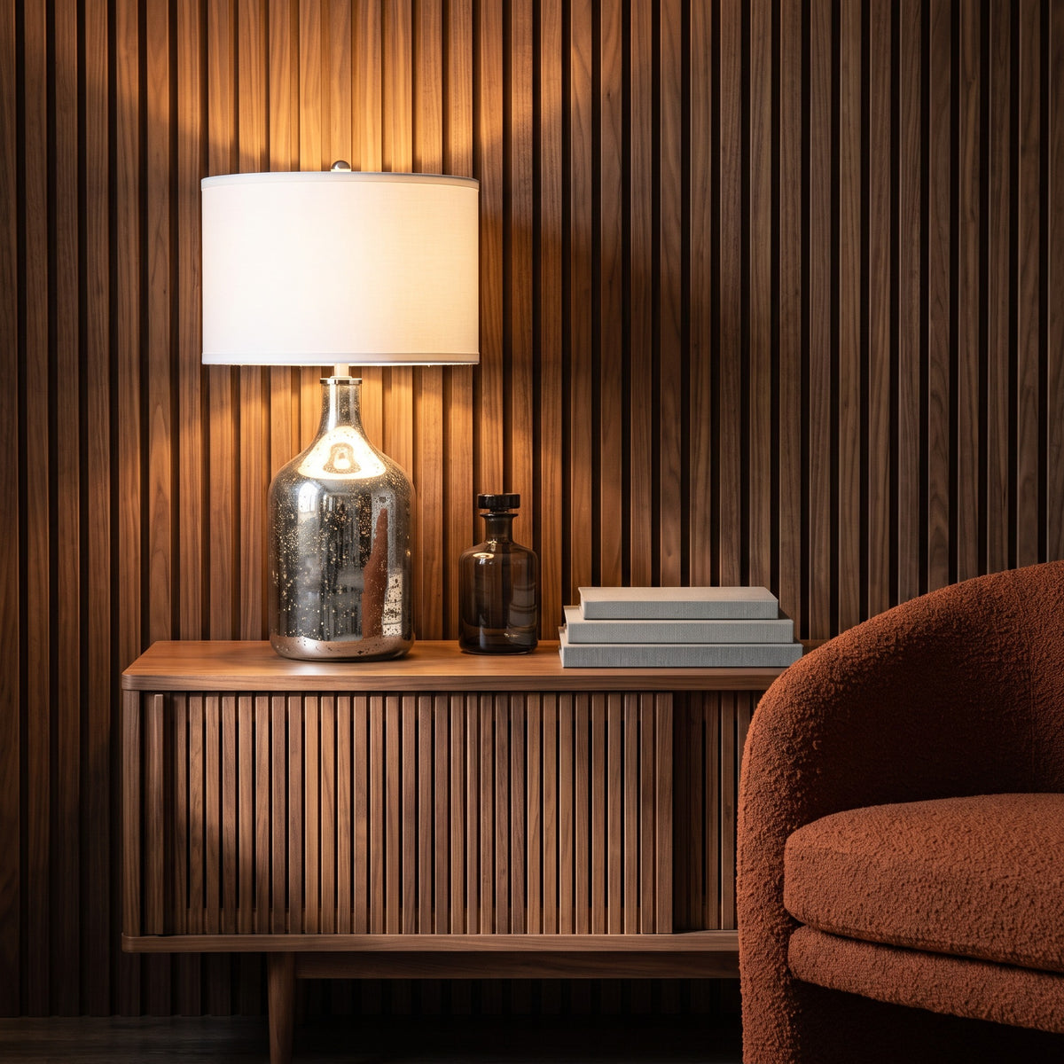 Mercury glass table lamp with white drum shade lit on a walnut credenza beside a rust boucle chair