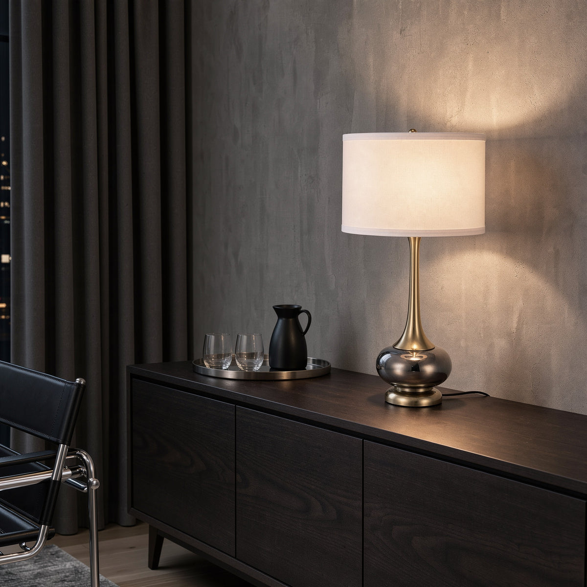 Gray glass and gold table lamp, lit, with white drum shade on a dark wood sideboard by a gray plaster wall