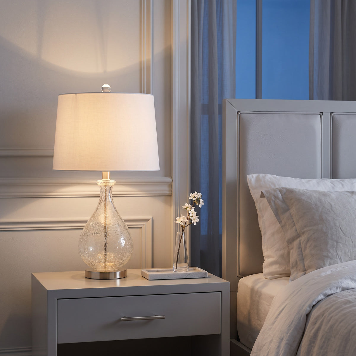 Crackle glass table lamp with white linen drum shade and nickel base lit on a white nightstand by a bed