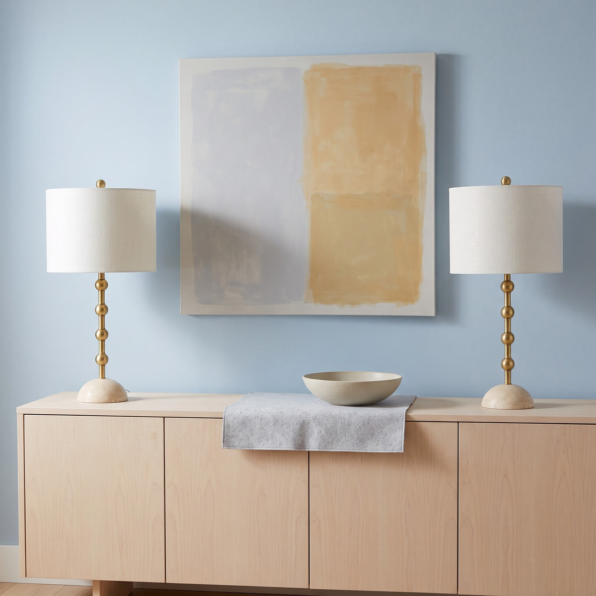 Pair of gold table lamps with travertine bases and off-white drum shades on a light wood sideboard