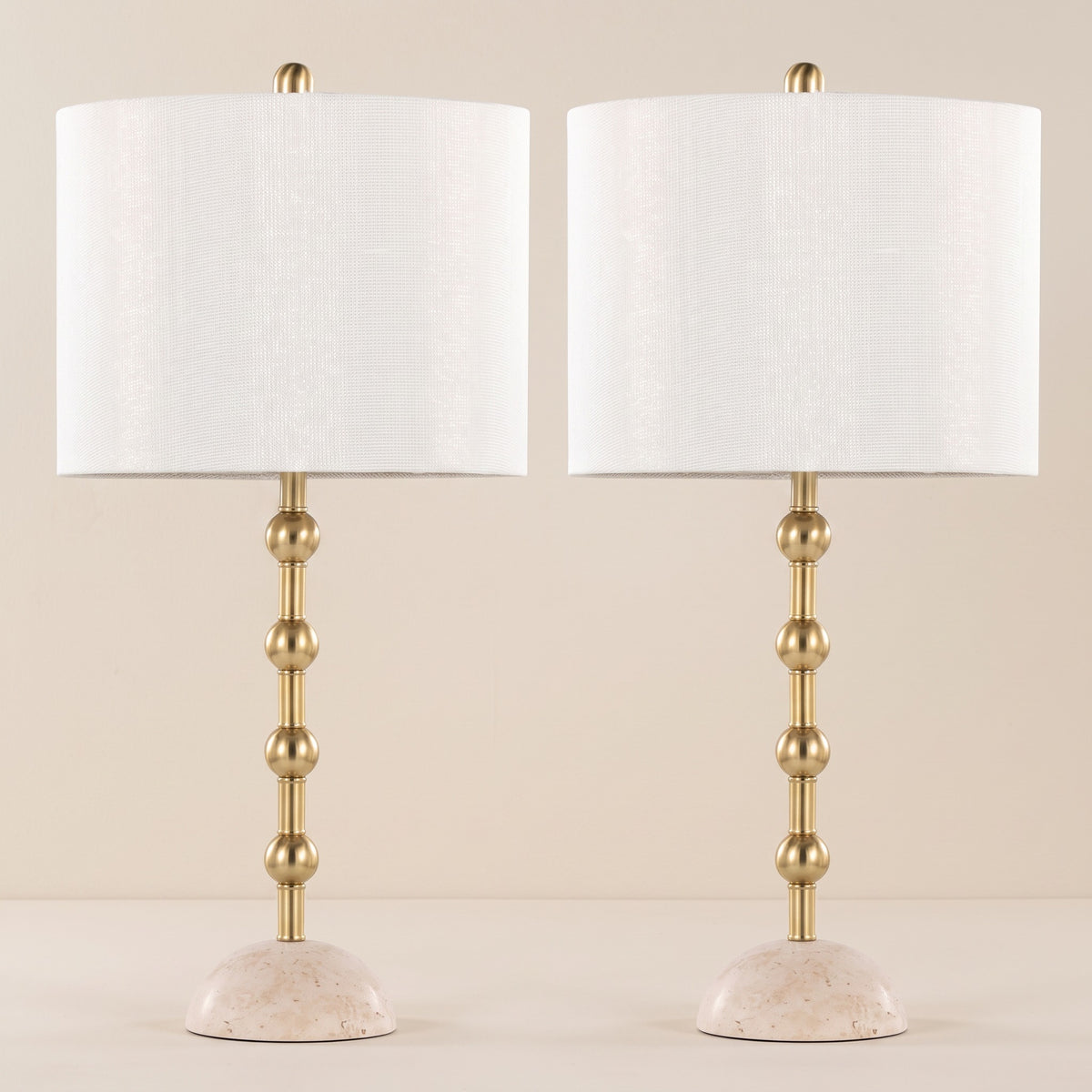 Pair of gold metal table lamps with stacked sphere stems, travertine bases and off-white shades on an ivory backdrop