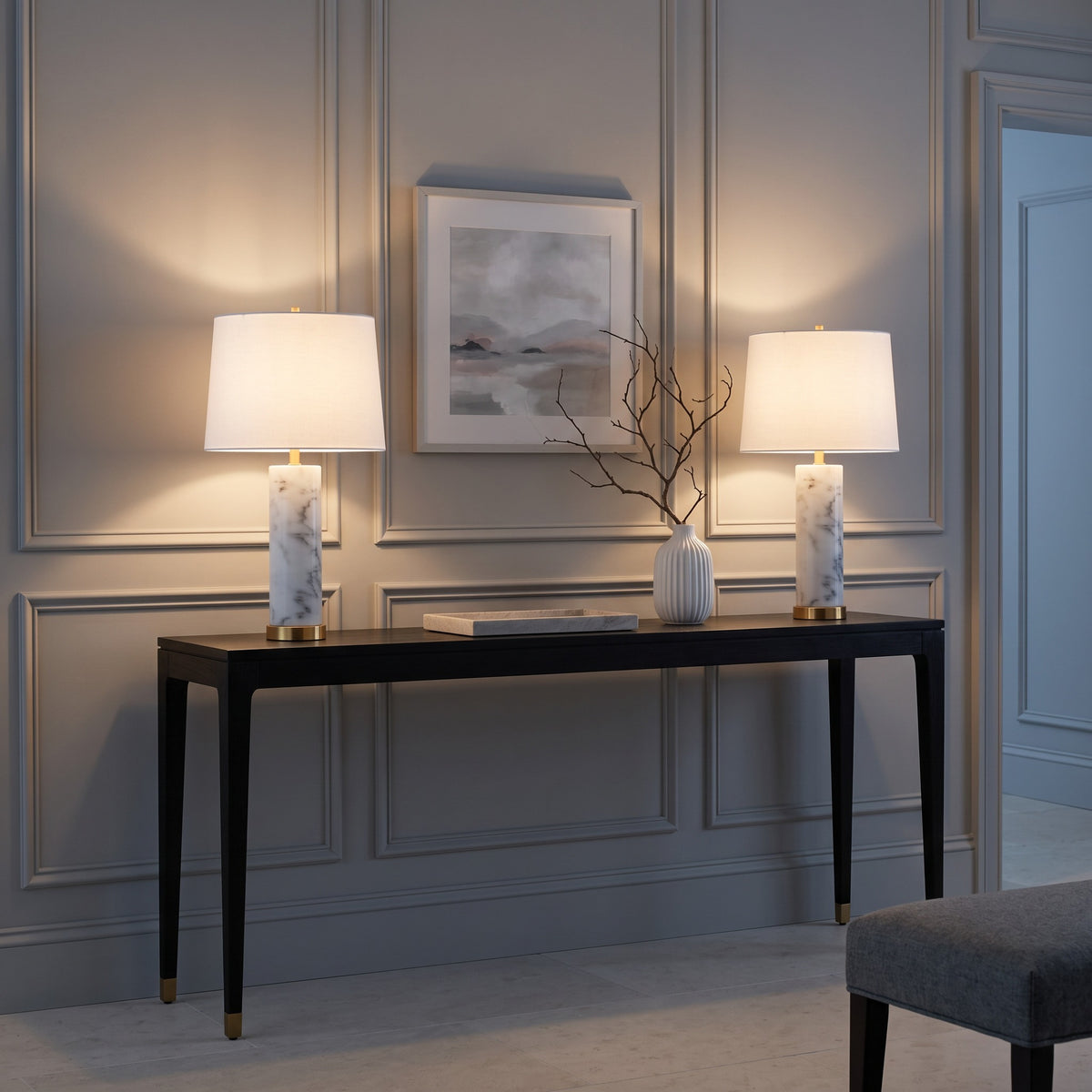 Pair of white marble table lamps with brass bases and white linen shades, lit on a black console table
