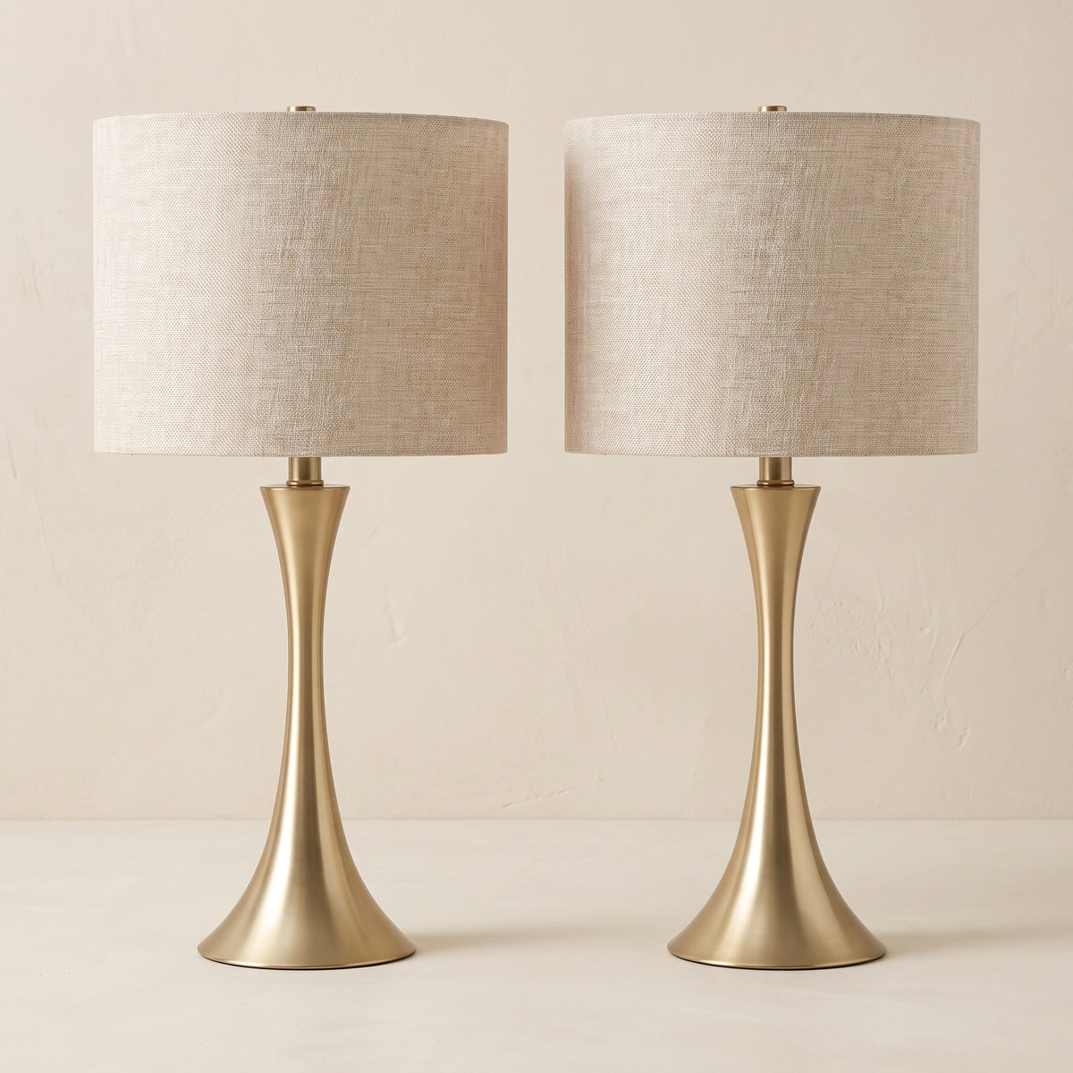Pair of gold table lamps with hourglass bases and linen-look drum shades on an ivory plaster backdrop
