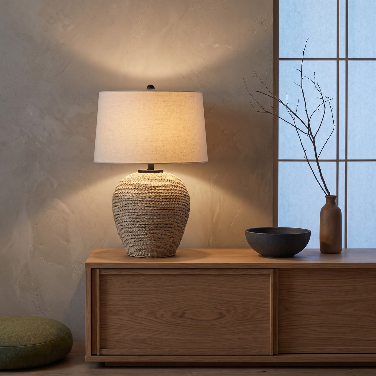 Rattan table lamp with beige linen drum shade lit on an oak console beside a shoji window