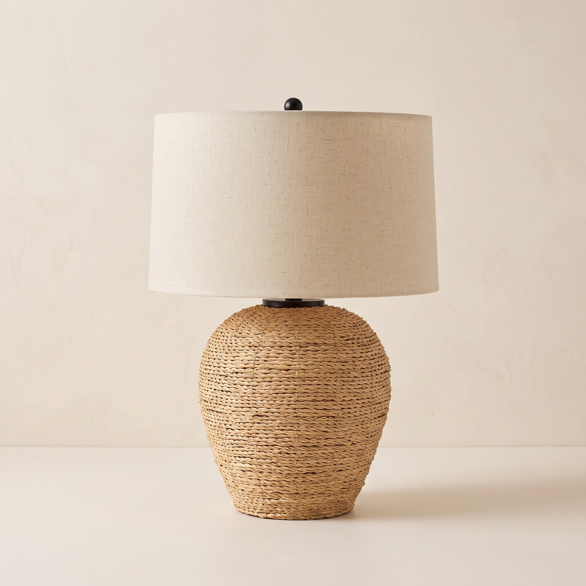 Rattan table lamp with ginger jar base, beige linen drum shade and black finial on an ivory backdrop