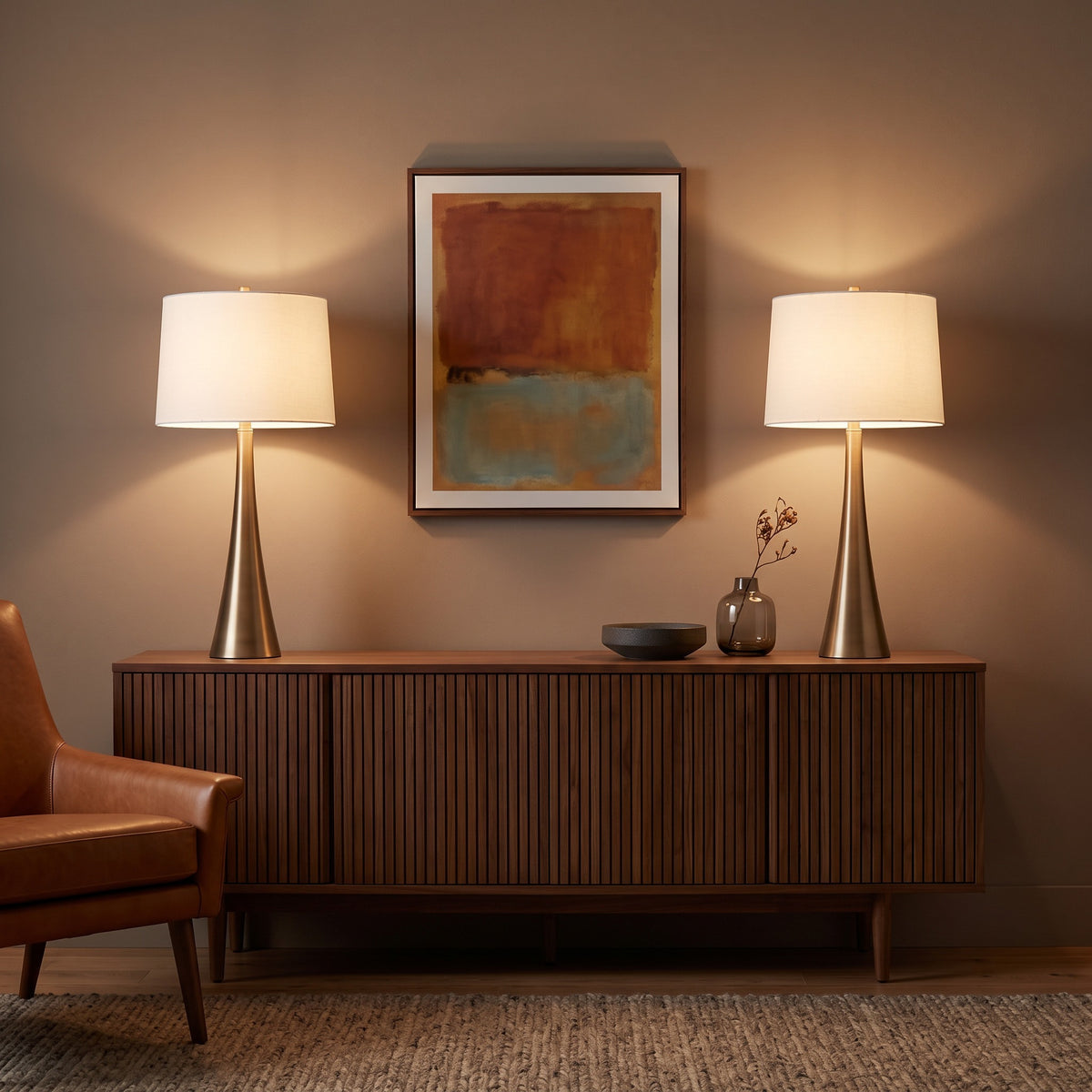 Pair of bronze table lamps with white linen shades lit on a walnut slatted credenza beside a leather armchair