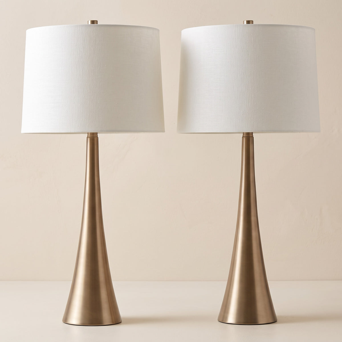Set of two bronze finish table lamps with tapered bases and white linen shades on an ivory plaster backdrop