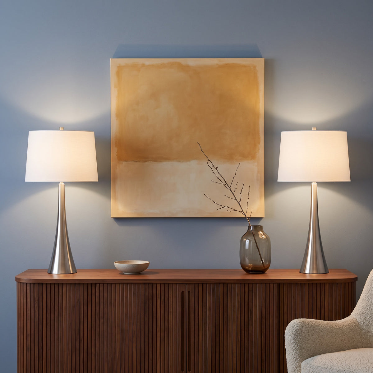 Pair of brushed nickel table lamps with white linen shades, lit, on a fluted walnut console against a blue wall