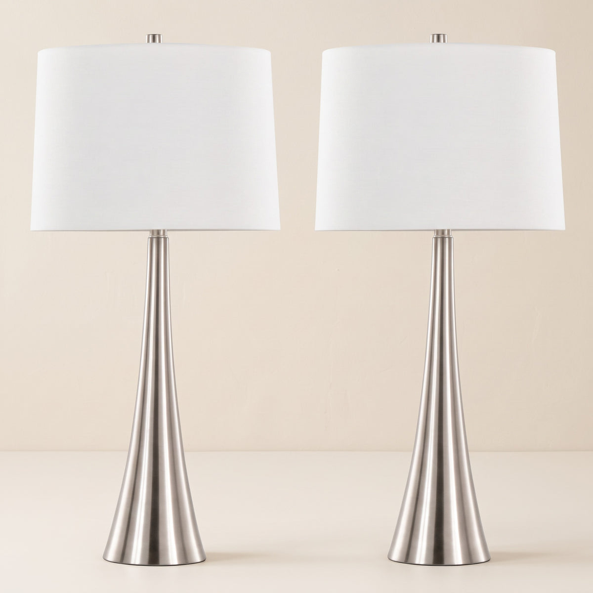 Pair of brushed nickel table lamps with white linen shades on an ivory plaster backdrop