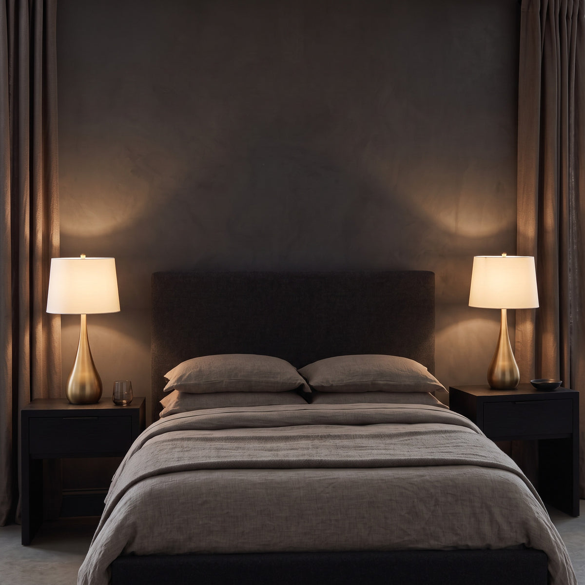 Pair of gold teardrop table lamps with white linen shades lit on black nightstands beside a bed