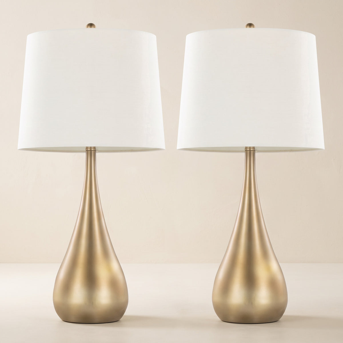 Pair of gold teardrop table lamps with white linen drum shades on an ivory plaster backdrop