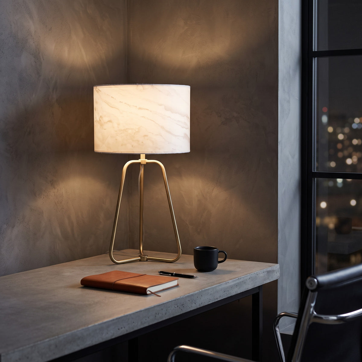 White marble-look drum shade table lamp on a brass finish tripod base, lit on a concrete desk by a night window
