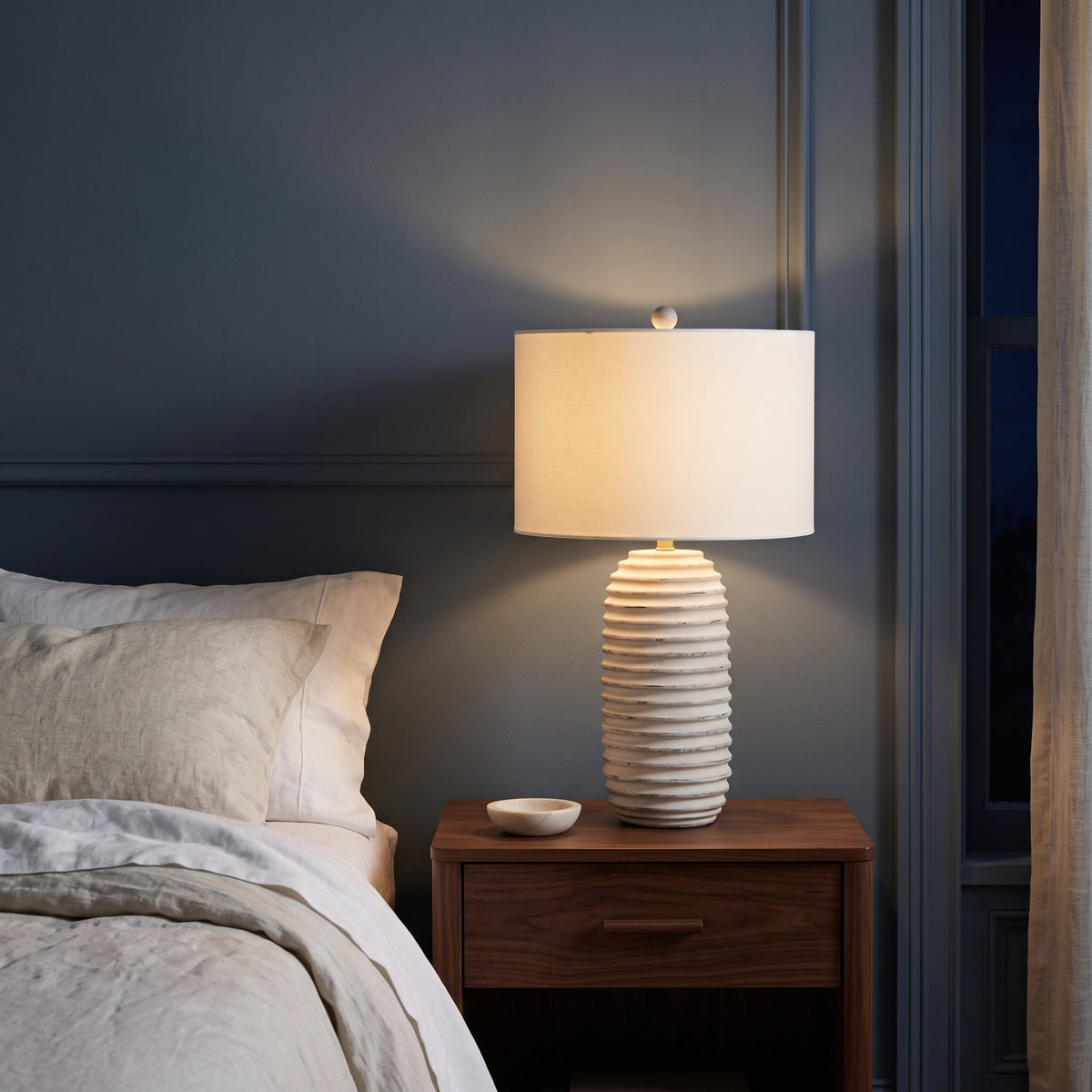 Cream ribbed table lamp with ivory drum shade, lit, on a walnut nightstand beside a bed