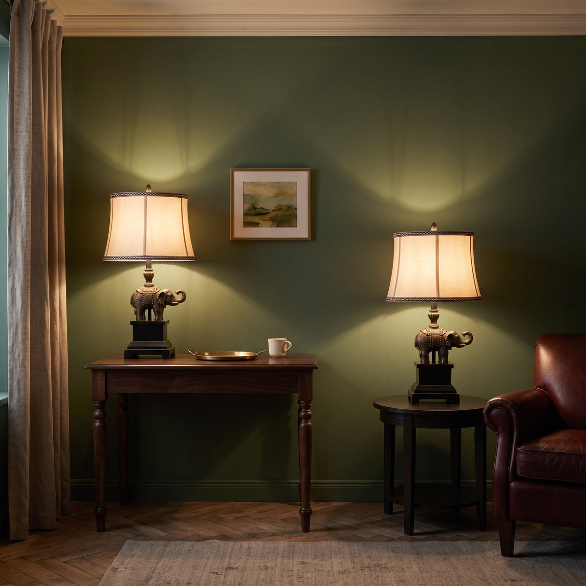 Pair of bronze elephant table lamps with linen shades lit on a walnut console and side table by a green wall