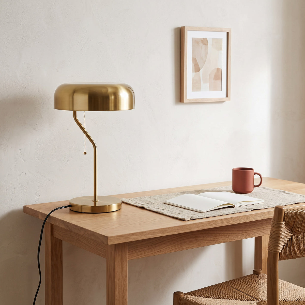 Gold metal table lamp with dome shade and pull chain on an oak desk beside a mug and open notebook