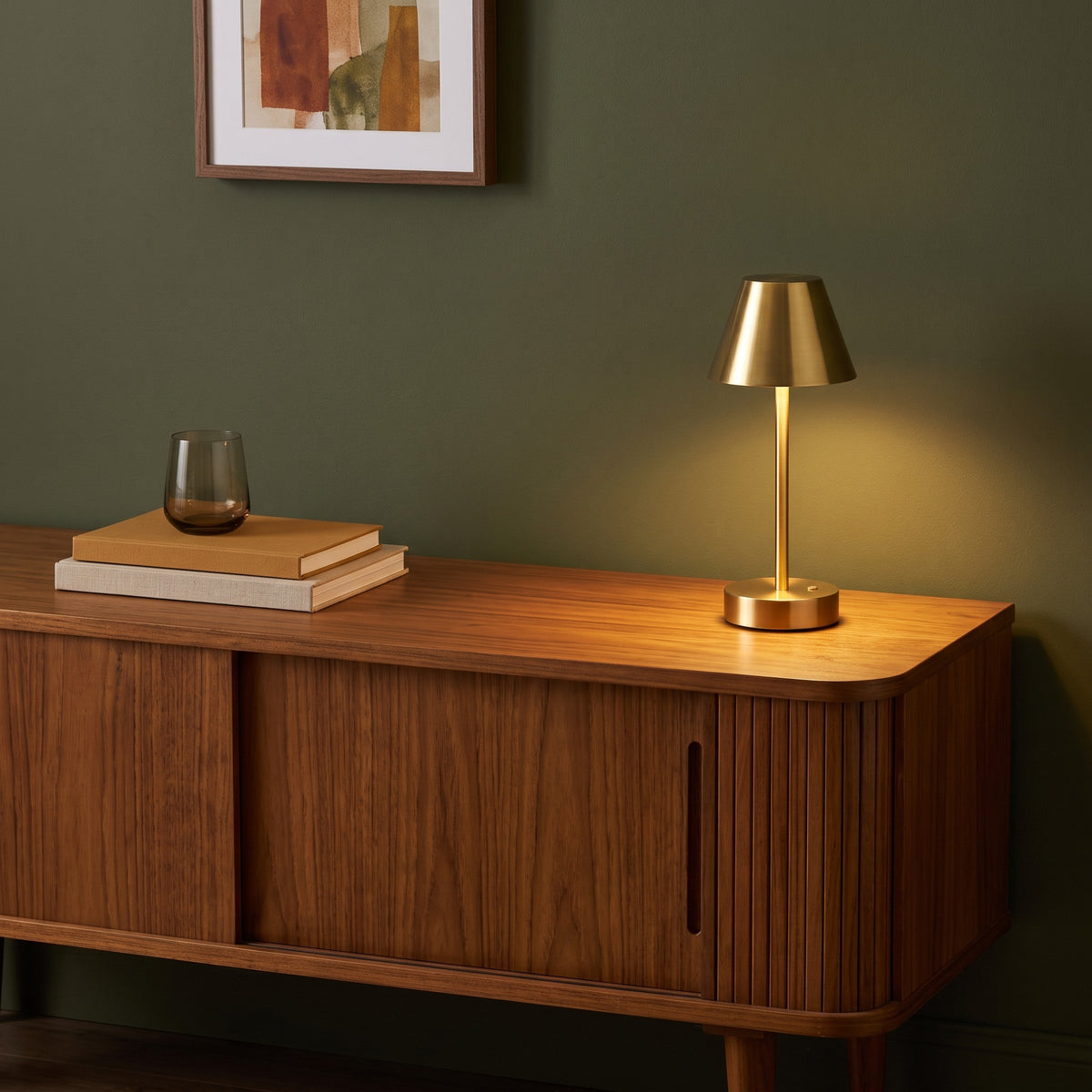 Brass cordless table lamp with cone shade glowing on a walnut sideboard against a green wall