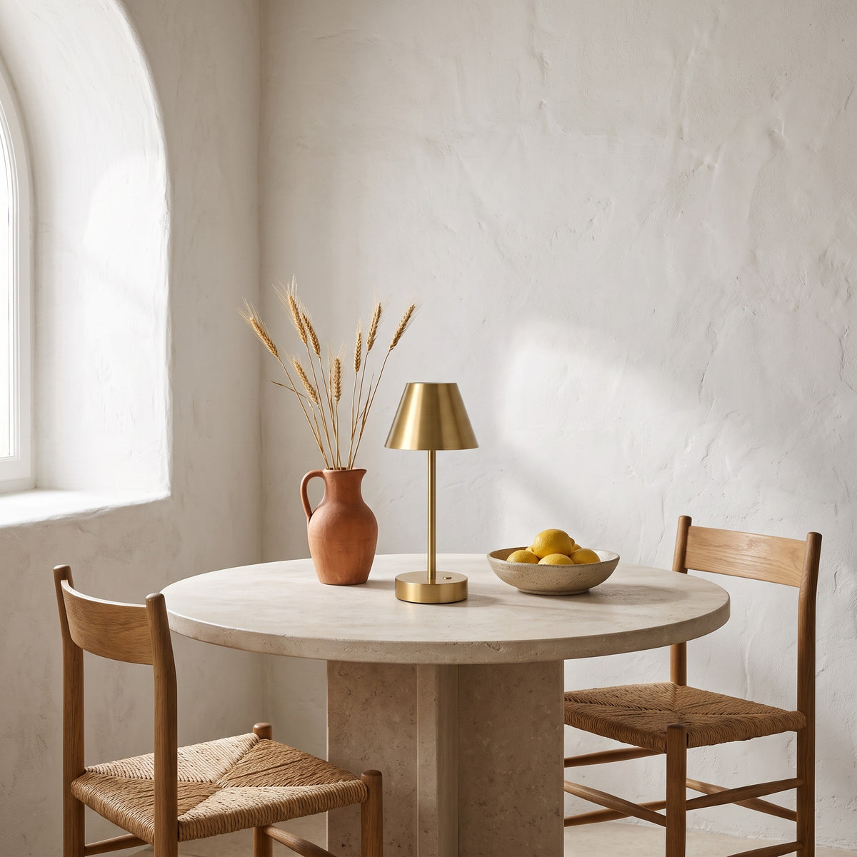 Brass finish cordless table lamp with cone shade on a round stone-look dining table beside a clay pitcher and lemons