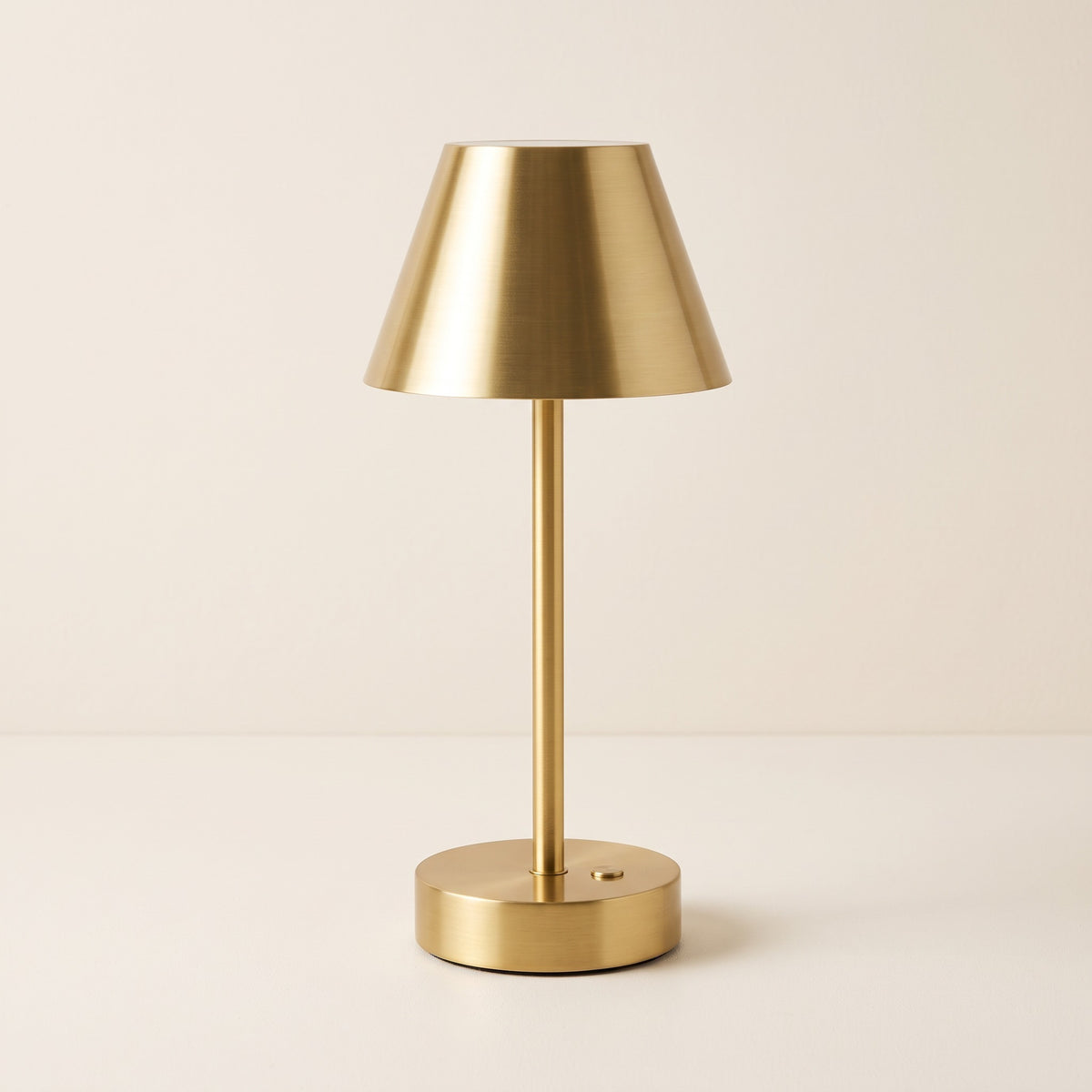 Brass finish cordless table lamp with cone shade, slim stem and round base on an ivory backdrop